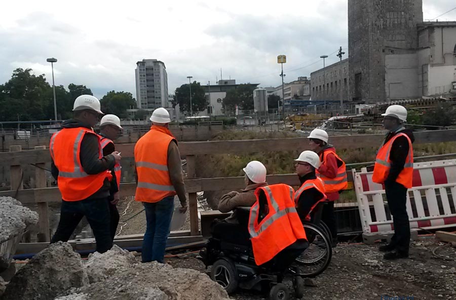 7 Personen mit Schutzhelm und Warnweste auf der Baustelle, davon zwei im Rollstuhl